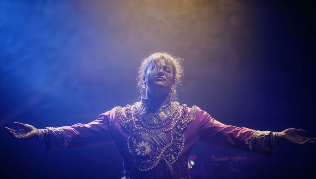 Singer raising arms on stage wearing beaded gown and layered jewelry under amber-blue lights