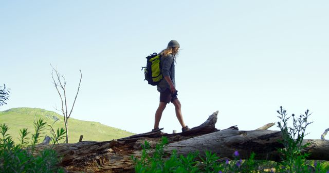 Young woman wearing a backpack, walking on a sizable fallen tree trunk in a lush forest or park in summer. Ideal for content related to outdoor adventures, hiking experiences, travel blogs, fitness activities, and nature exploration.