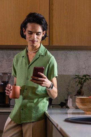 Asian man leaning on kitchen counter holding mug and checking smartphone in green shirt