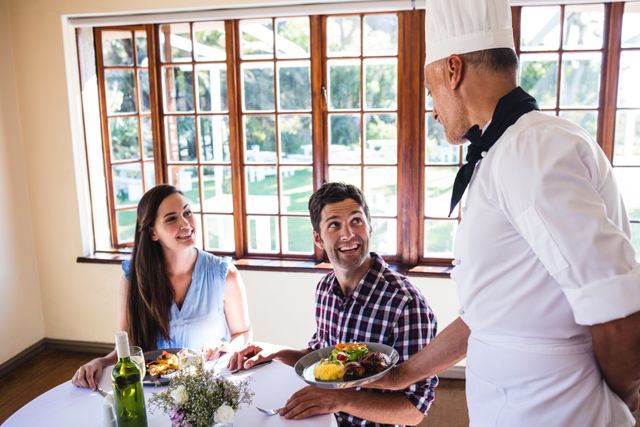 Chef presenting a gourmet meal to a young couple seated at a restaurant table. Ideal for use in advertisements for restaurants, culinary schools, hospitality services, and dining experiences. Highlights themes of fine dining, customer service, and romantic dining.