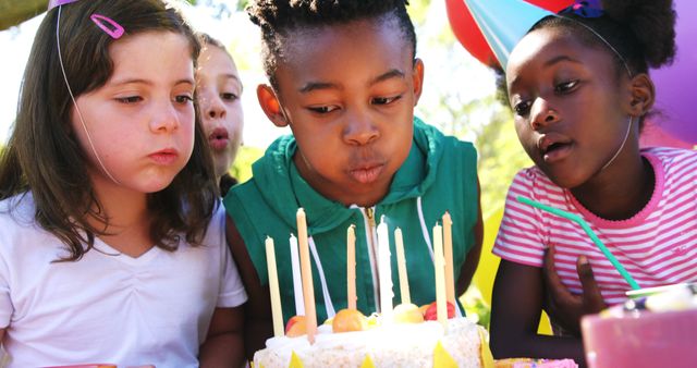 A diverse group of children is gathered around a birthday cake, with a young African American girl preparing to blow out the candles, with copy space. The image captures a moment of celebration and excitement during a child's birthday party.
