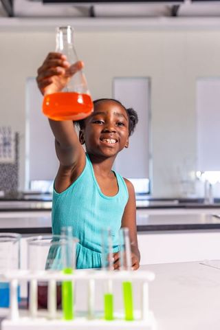 African american girl holding orange flask smiling in stem lab with bright test tubes