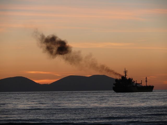 Cargo ship emitting smoke in tranquil ocean at sunset with mountain silhouettes in the background. Useful for illustrating themes related to maritime transportation, environmental pollution, industrial impact on nature, and picturesque evening seascapes.