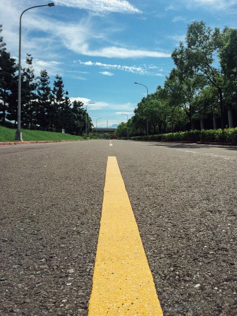 The image shows an empty road taken from a low perspective, focusing on the yellow line in the center. The scene is set under a clear blue sky with scattered clouds, bordered by green trees on both sides. Ideal for representing tranquility, freedom, or the concept of an open journey. Useful for travel, motivational, and urban design projects.