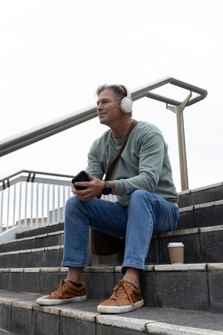 Middle-aged man relaxing on urban steps listening to music with coffee and phone