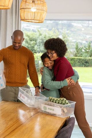 Black family hugging while sorting paper and glass recycling bins in dining room