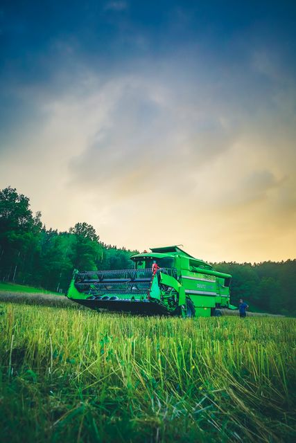 This image features a green combine harvester working in a field as the sun sets, casting beautiful colors in the sky. The lush, green machine stands out against the golden tones of the crops and the backdrop of trees and dusk. This image can be used for agricultural themed presentations, farming and machinery advertisements, or to depict rural life and productivity.