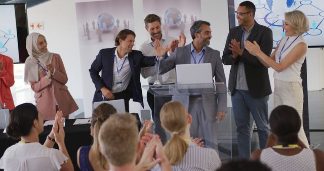 Group of business professionals from diverse backgrounds celebrating a successful presentation in a conference hall. They are standing at a podium while the audience applauds. Suitable for use in articles and advertisements related to corporate events, team achievements, diversity in the workplace, and professional success.