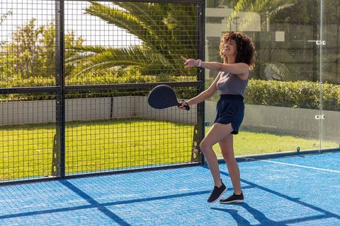 Smiling african woman playing padel on blue court pointing in sunny outdoor club