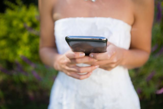 Bride in white dress using mobile phone outdoors in garden. Ideal for wedding planning, technology use in weddings, social media communication, modern bride lifestyle, and outdoor wedding themes.