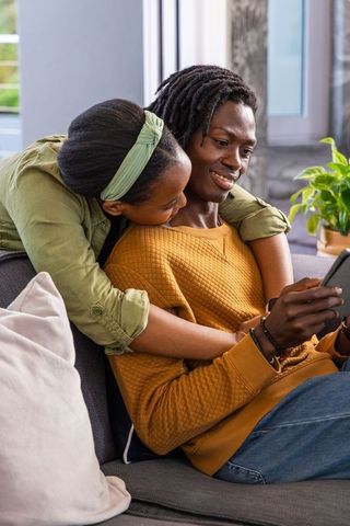 African American couple cuddling on sofa, sharing phone and smiling in cozy living room