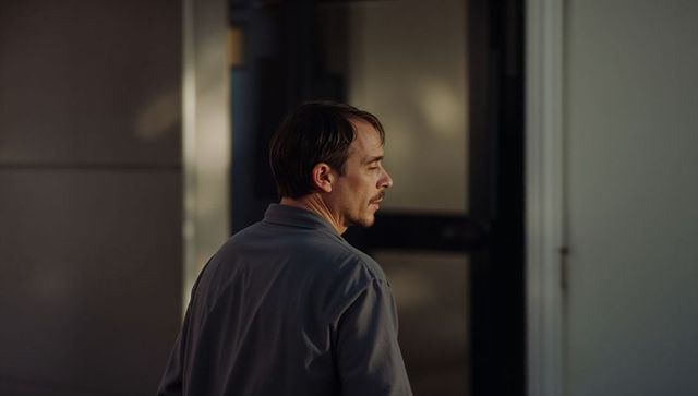 Moody sidelit profile of man turning head in dim hallway wearing gray collared jacket