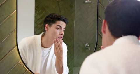 Young man examining skin in bathroom for self-care routine