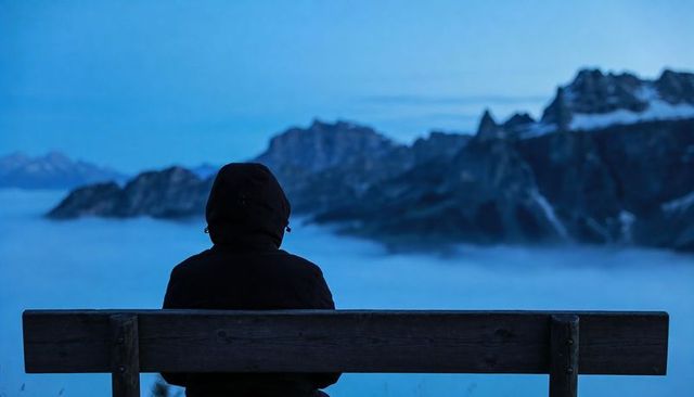 Solitary hiker sitting on bench watching fog-filled alpine valley at blue hour silhouette