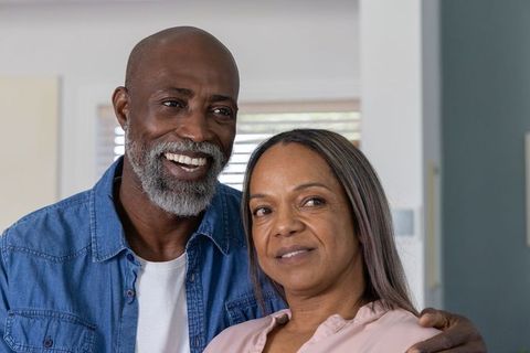 Senior couple smiling and embracing at home, man resting arm on partner's shoulder