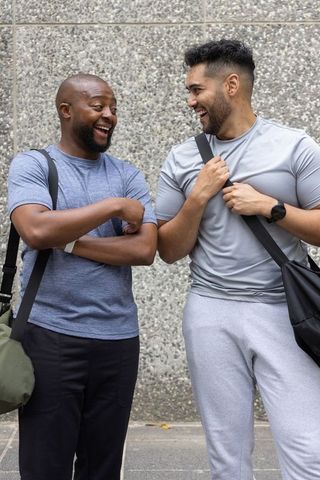 Two friends laughing and chatting after workout on urban sidewalk carrying duffel bags