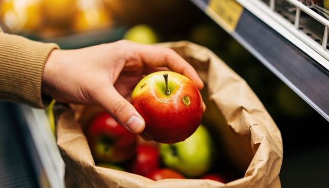 Hand picking red-yellow apple with small bruise from paper bag at grocery produce display