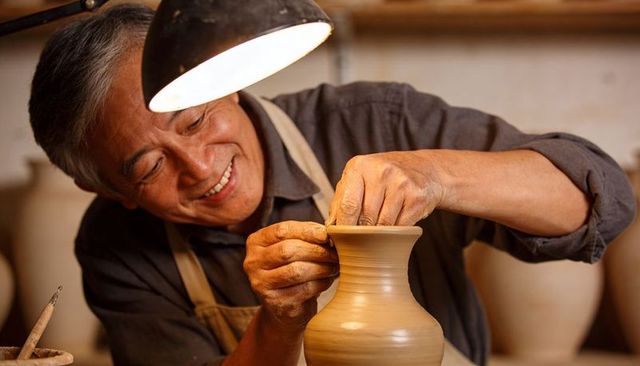 Skilled potter shaping clay vase under lamp, smiling during handmade ceramics craft