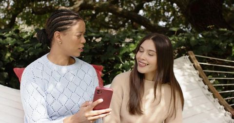Diverse women relaxing on hammock in garden sharing smartphone, smiling and chatting
