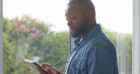 African man standing by window checking tablet and watch wearing denim shirt lifestyle