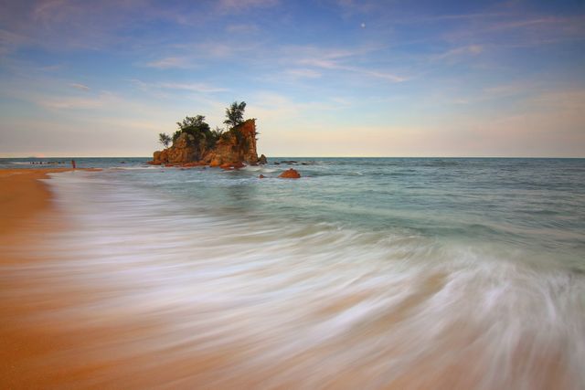 Long exposure of a serene beach at sunset with smooth waves washing onto sandy shoreline, small island with trees in the distance. Ideal for travel websites, nature blogs, relaxation content, backgrounds, greeting cards highlighting tranquility, beauty of coastal landscapes, perfect for illustrating peaceful retreats, beach vacations.