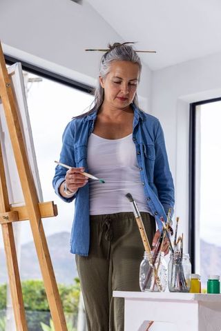 Senior woman painting in sunlit studio wearing denim shirt holding brush by easel