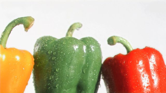 Three bell peppers – orange, green, and red – being soaked with water droplets against a white background. Perfect for use in advertising for fresh produce, healthy eating campaigns, farmers' markets, grocery stores, or cooking blogs focused on vegan or vegetarian recipes.