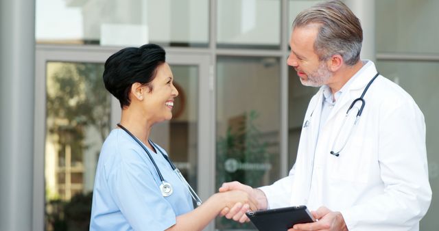 Healthcare professionals shaking hands outside a hospital, symbolizing collaboration and teamwork. Nurse in blue scrubs and doctor in a white coat smile while greeting each other. Perfect for themes related to medical cooperation, professional respect, hospital environments, healthcare industry, and teamwork in healthcare settings.