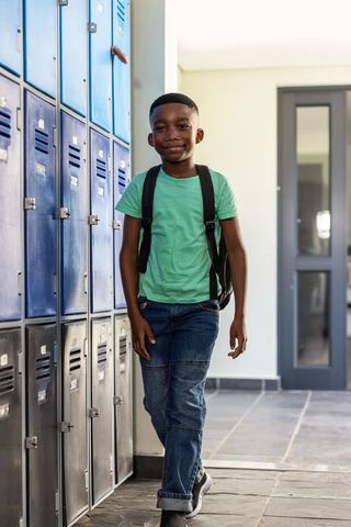 Preteen african american boy walking school hallway with backpack by blue lockers smiling