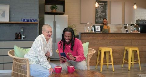 Smiling diverse friends leaning in sharing smartphone over lattes in cozy cafe lounge