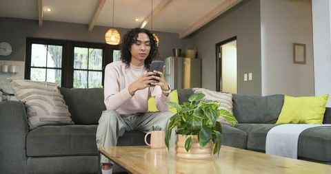 Non-binary adult using smartphone on modern grey sofa in cozy open-plan living room