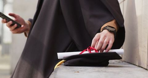 Graduate checking phone while holding mortarboard and diploma on stone bench