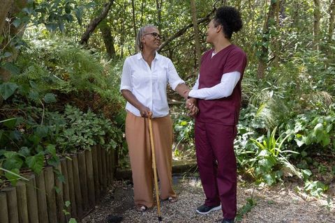 Caregiver supporting senior woman walking with cane on garden path, outdoor elder care