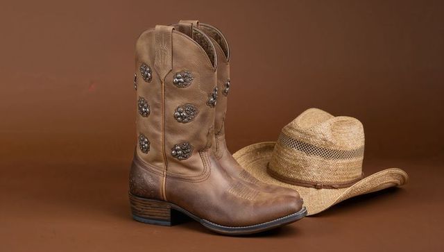 Studded leather cowboy boots and straw hat on earthy brown background