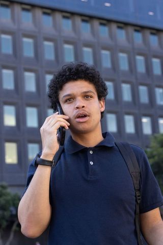 Young african american commuter talking on smartphone in urban plaza wearing navy polo and backpack