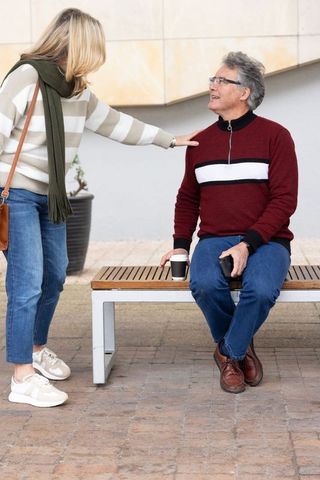 Mature couple chatting on bench, man holding coffee and phone, woman touching shoulder