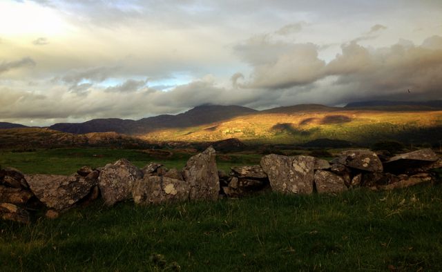 This image showcases a dramatic landscape during the golden hour. The foreground features a rugged stone wall, while the lush green fields stretch towards the horizon. The mountains in the background are bathed in a warm golden light under a partly cloudy sky, creating a picturesque scene. This image is ideal for nature-themed projects, travel brochures, or environmental campaigns.
