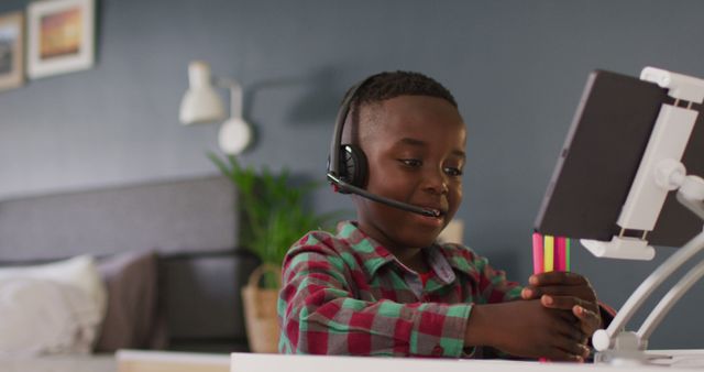 Young boy using digital tablet with headset, participating in online learning session from home. Ideal visual for content related to remote education, digital communication, virtual classrooms, and child technology engagement.