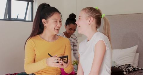 Sharing makeup palette and laughing friends sitting on bed in cozy bright bedroom scene