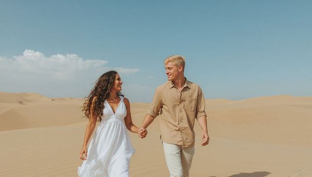 Couple walking hand-in-hand across sunlit desert dunes, flowing white dress, beige shirt