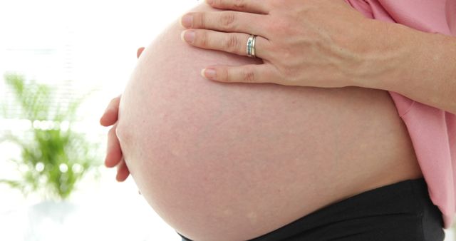 Close-up of pregnant woman gently holding her bare belly with hand, standing at home. This image can be used for content related to maternity, prenatal care, pregnancy health, motherhood, and family planning.