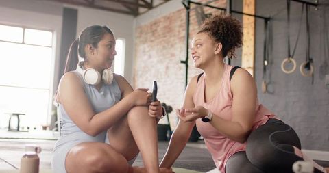 Diverse women chatting on exercise mats at functional fitness gym with smartphone