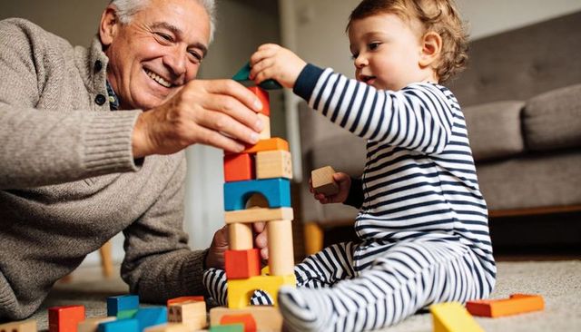 Grandfather building colorful wooden block tower with toddler on living room carpet