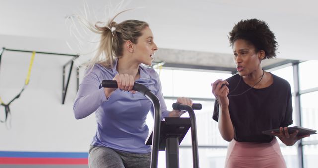 Image of diverse female fitness trainer with stopwatch and woman on exercise bike working out at gym. Exercise, fitness and healthy lifestyle.