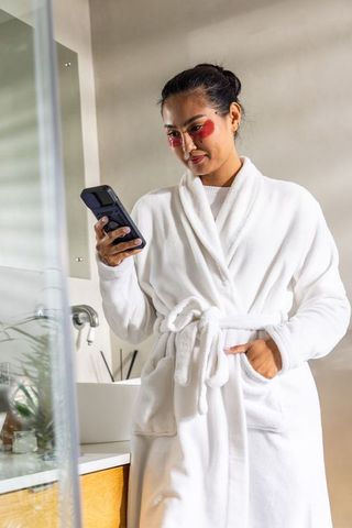 Asian woman wearing white robe checking smartphone with red under-eye patches