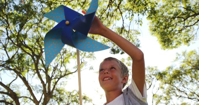 Young boy enjoys playing with a blue pinwheel under the trees on a bright summer day. Great for depicting themes of childhood joy, outdoor activities, summer fun, and carefree play. Can be used for websites, educational materials, or advertising children's toys and outdoor products.