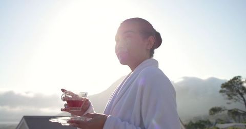 Mid-adult asian woman relaxing on terrace in white bathrobe holding cup of tea at sunrise