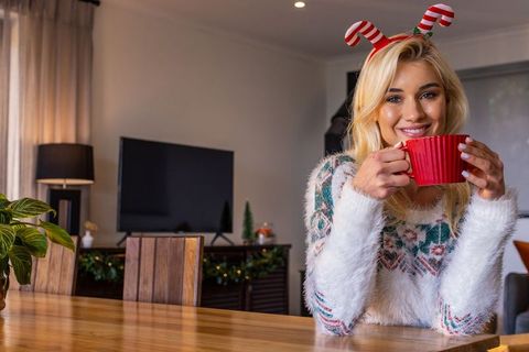 Smiling woman sipping hot cocoa wearing cozy holiday sweater and candy cane headband