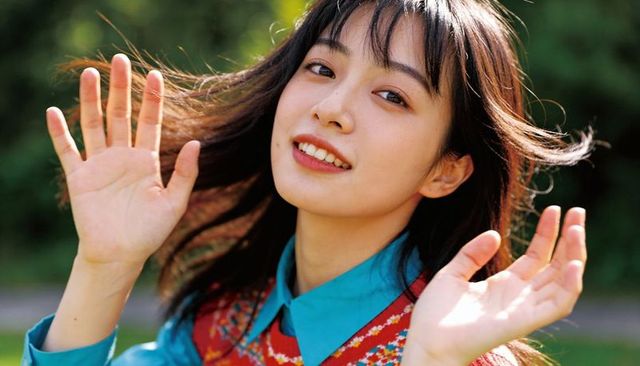Smiling asian woman raising hands in park wearing turquoise shirt and patterned vest