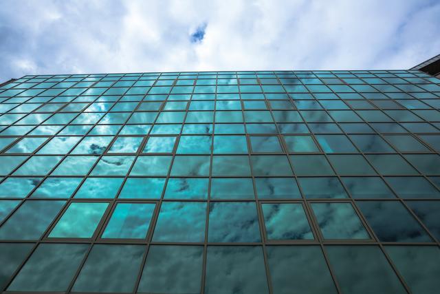 Low angle view of a modern glass skyscraper reflecting the blue sky and clouds. Ideal for use in corporate presentations, real estate marketing, architectural portfolios, and urban development projects.